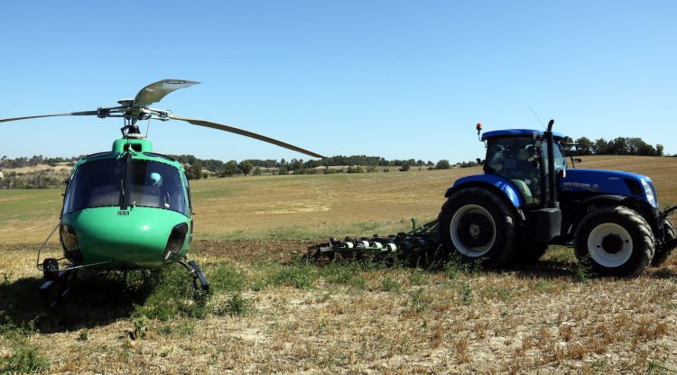 Un tractor llaurant un camp de cereal ja segat al costat de l'helicòpter dels Agents Rurals. ACN