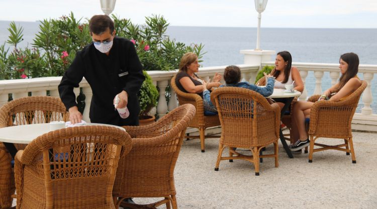 Un cambrer desinfectant les cadires de la terrassa de l'Hotel Costa Brava de Platja d'Aro amb unes dones prenent un cafè al darrera. ACN